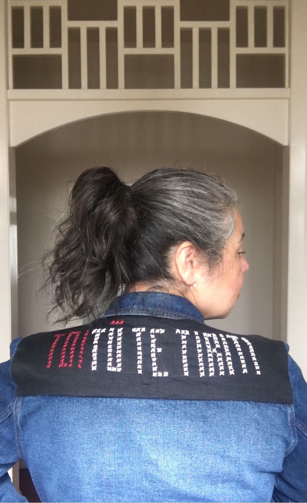 Photo of a middle aged wahine Māori wearing a denim jacket with a “TOITŪ TE TIRITI” cross stitched patch across the shoulders.