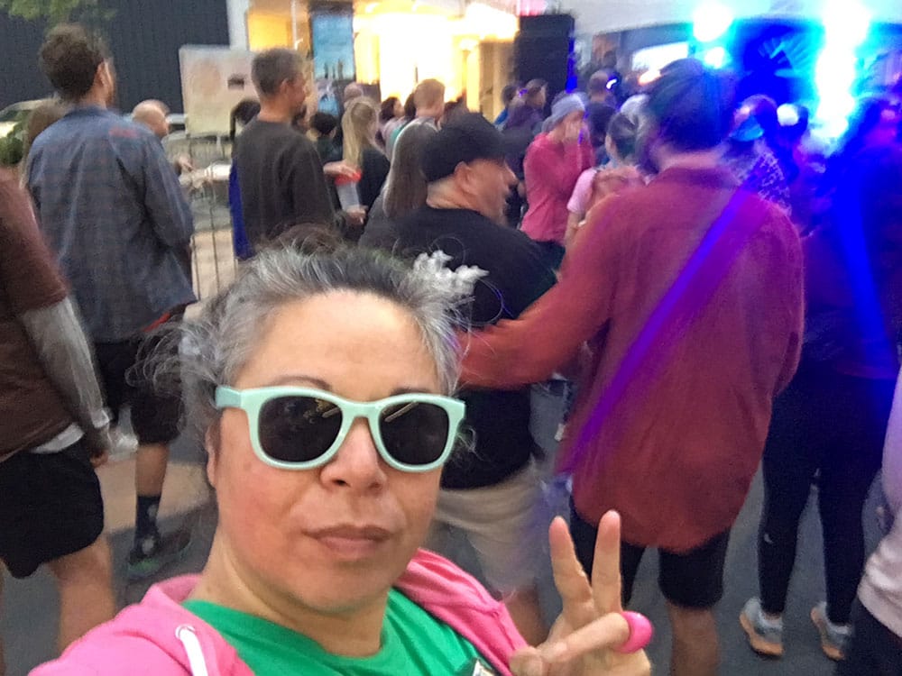 A grey haired woman in sunglasses, a green tshirt and pink hoodie throws up a peace sign in a crowd of ravers.