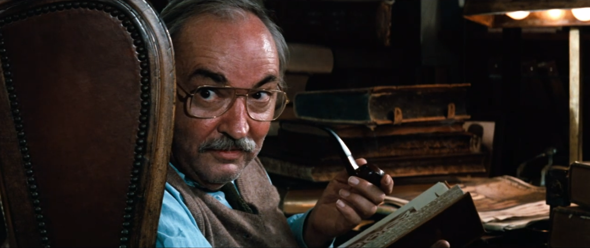 Screengrab of a moustachioed balding man in sex offender spectacles with a book and pipe in hand.