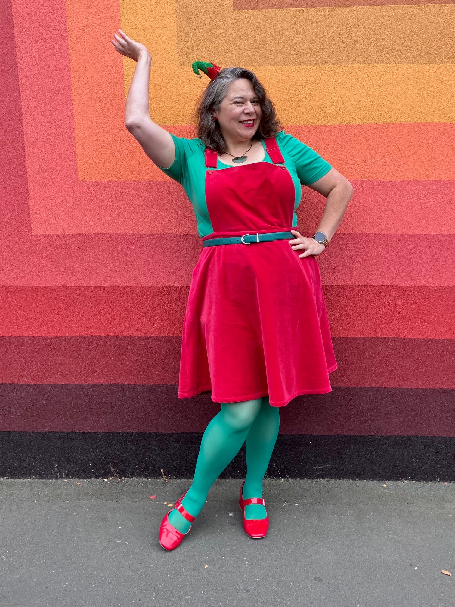 Moata posing in a red velveteen pinafore, green top, green belt, green tights and red shoes. Plus teeny elf hat on head.