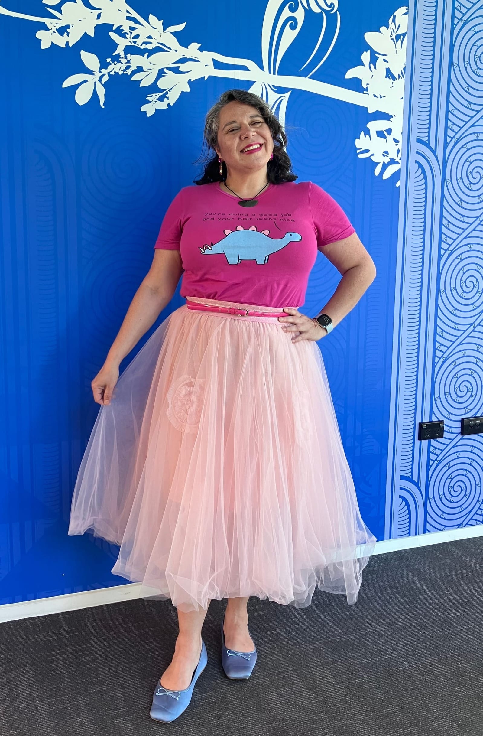 Moata looking pleased as punch with her life choices in a midi-length pink tulle skirt, blue satin ballet flat, pink belt, and Pink and blue Dinosaur Therapy tshirt.