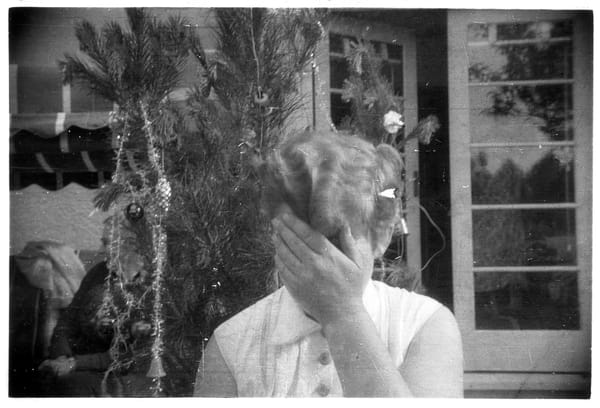 Photo of an unidentified person beside a Christmas tree hiding their face with their hand. Taken 1950s to 1960s.